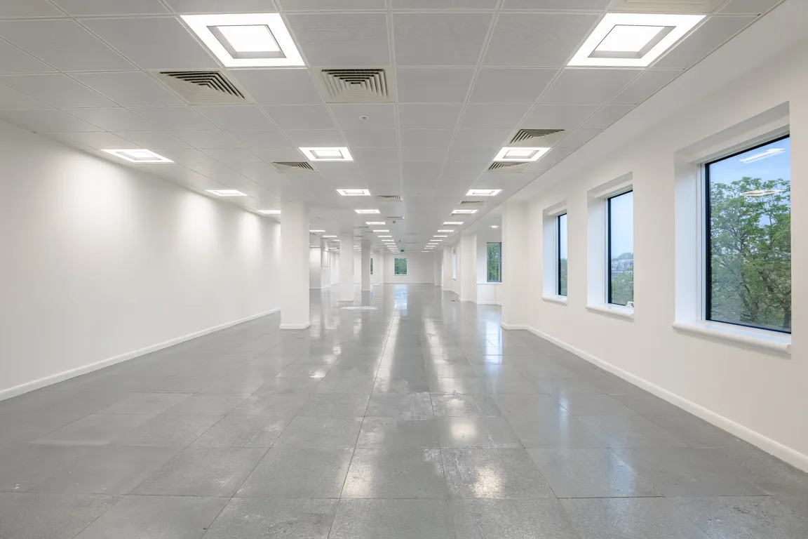 Empty, spacious office interior with white walls, grey tiled floor, ceiling lights, and windows showing trees outside.