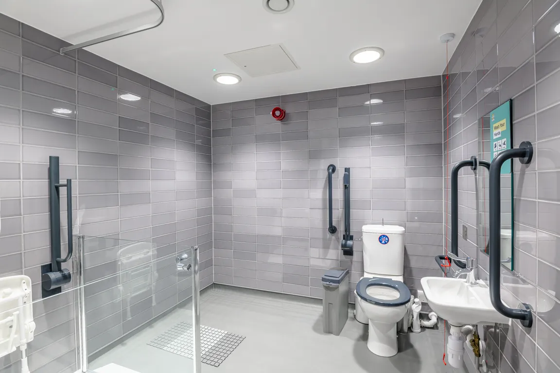 Accessible restroom with white toilet, grab bars, sink, foldable shower seat, and gray tiled walls.