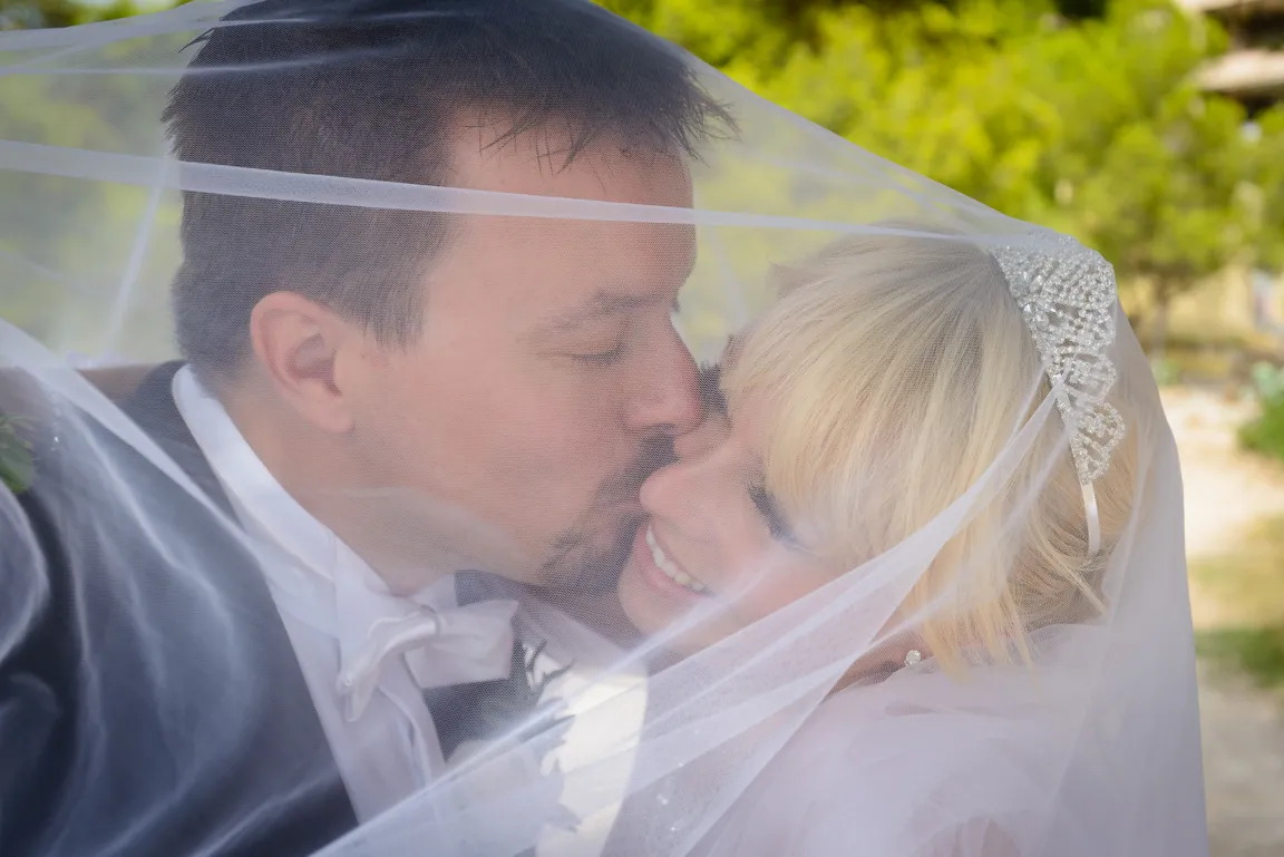Groom kissing bride on the cheek under her wedding veil with a blurred green background.