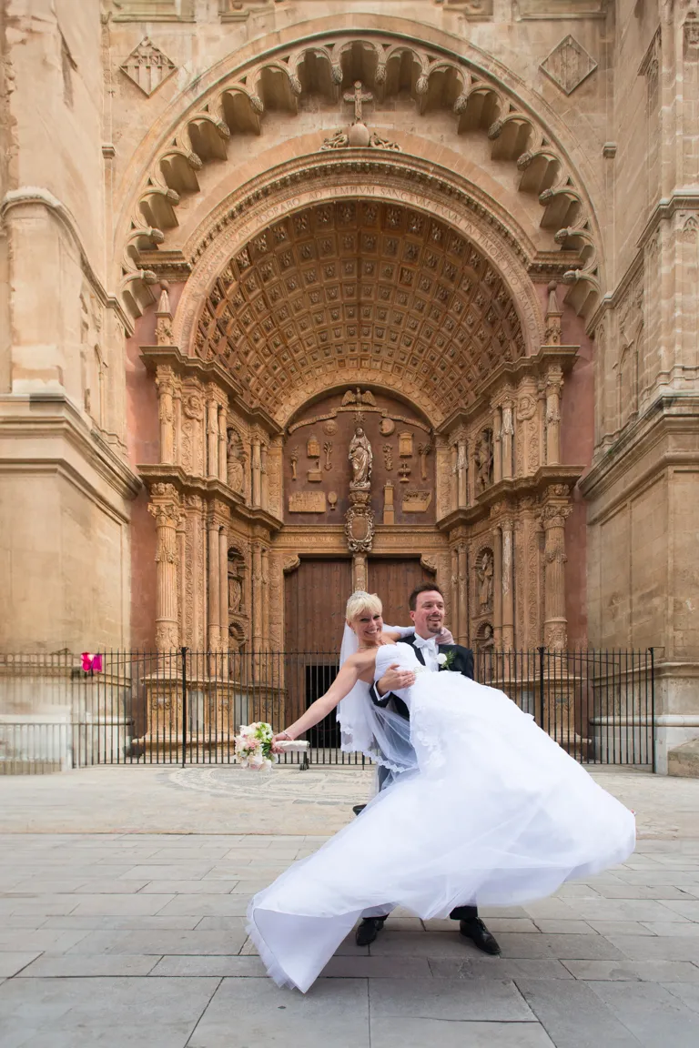 Groom in black tuxedo lifting bride in white wedding dress and veil in front of ornate cathedral entrance.