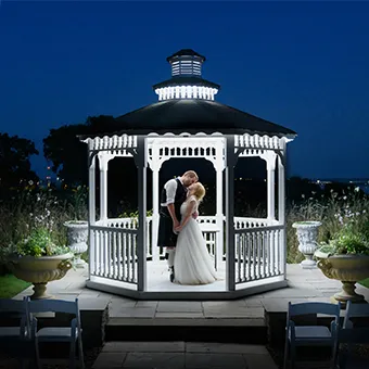 Bride and groom sharing a kiss inside a lit white gazebo at night.