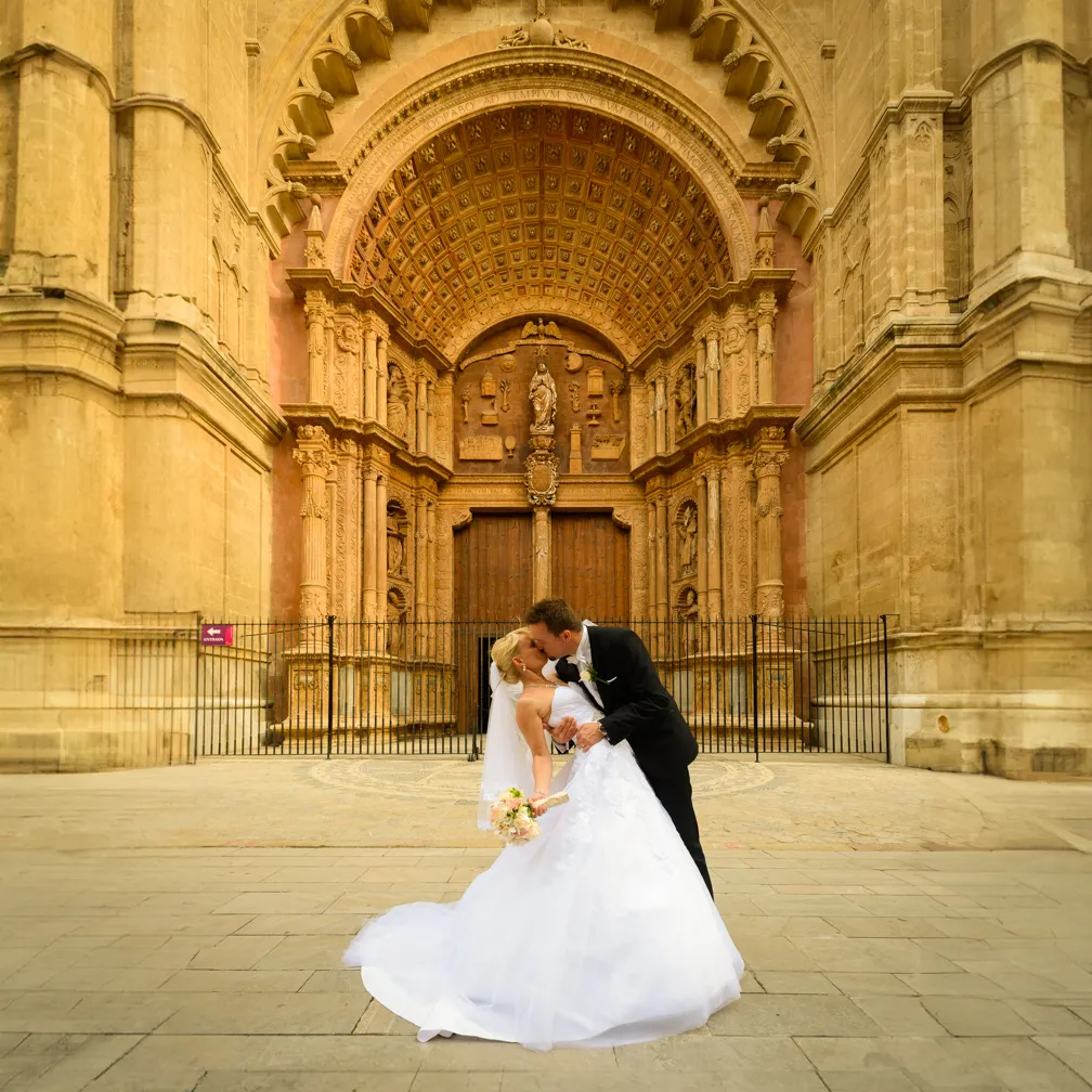 Bride and groom kissing in front of an ornate historic church entrance with stone carvings.