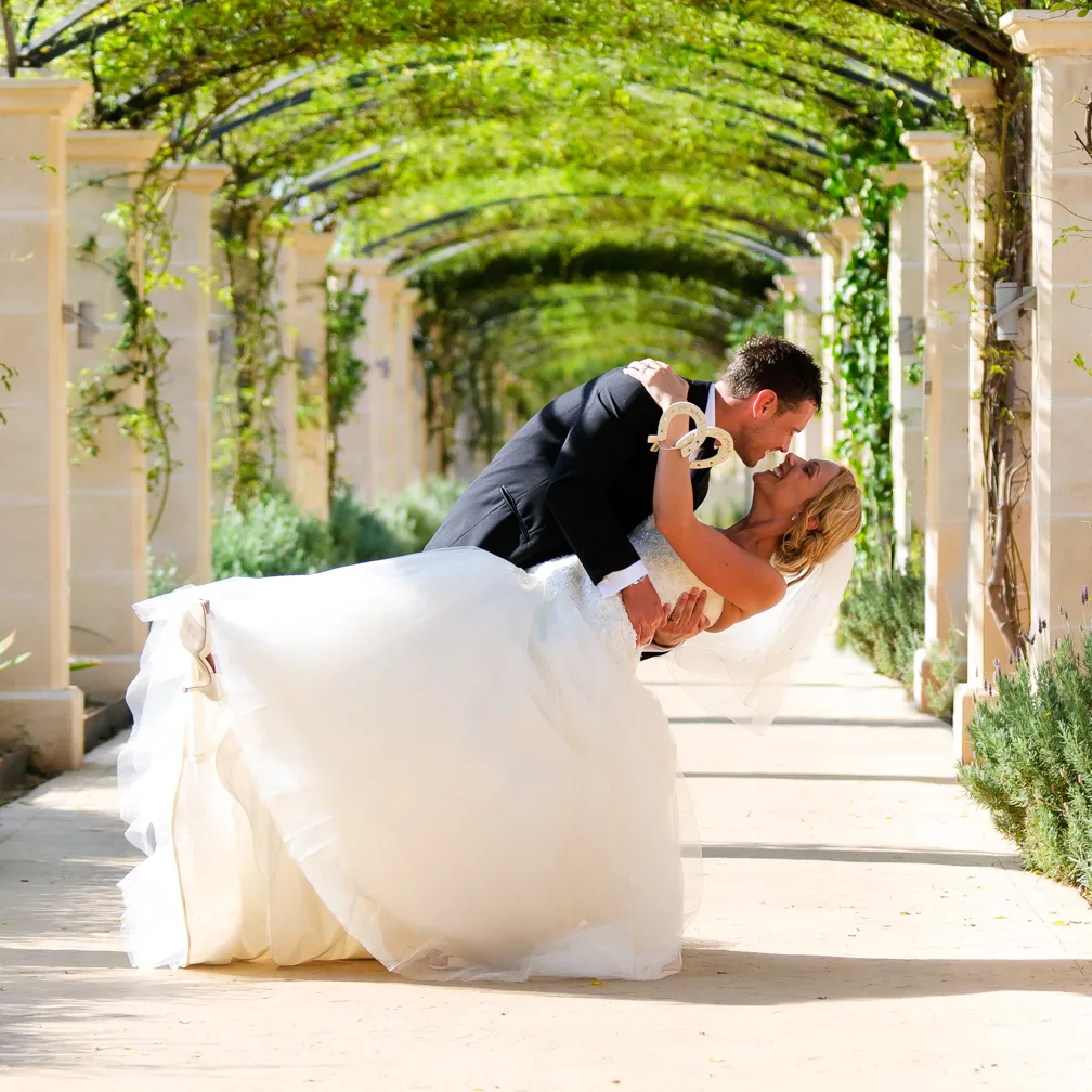 Bride and groom sharing a joyful embrace and kiss under a green vine-covered archway, the groom dipping the bride in her white wedding gown.