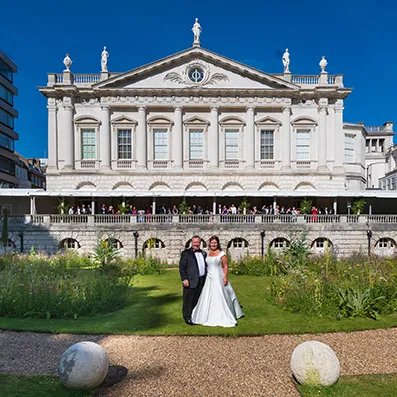 Bride and groom standing on a garden lawn in front of a grand historic building with statues on the rooftop and a clear blue sky.