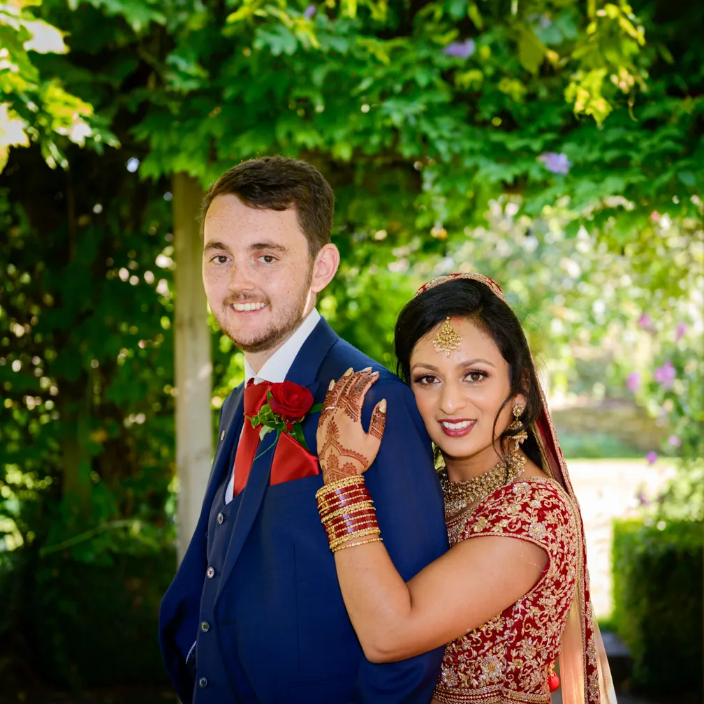 Bride in traditional red and gold attire with henna on her hand embraces groom in a navy blue suit with red rose boutonniere under green leafy canopy.
