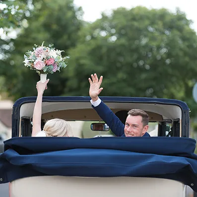 Newlywed couple waving and holding a bouquet while riding in the back of a vintage convertible car.