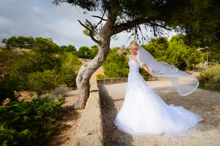 Bride in a white wedding dress and veil posing outdoors near a tree and stone ledge.