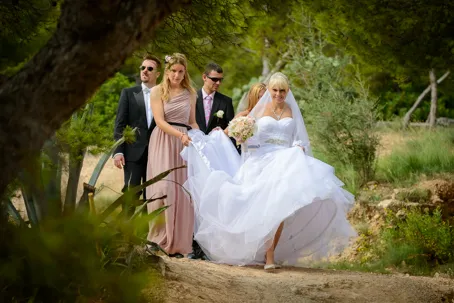 Bride in white wedding dress walking outdoors on a dirt path, accompanied by two men in suits and a woman in a pink dress.