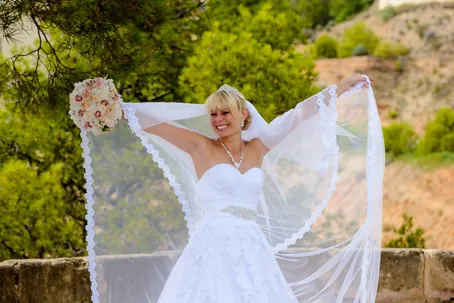 Smiling bride outdoors holding up her veil with one hand and a bouquet with the other against a green and hillside background.