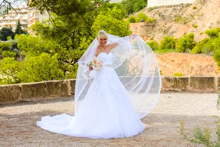Bride in a white wedding gown and veil holding a bouquet, smiling outdoors with trees and rocky hills in the background.