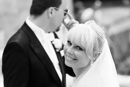 Smiling bride with veil and tiara looking at the camera, groom in suit wearing sunglasses in the background.