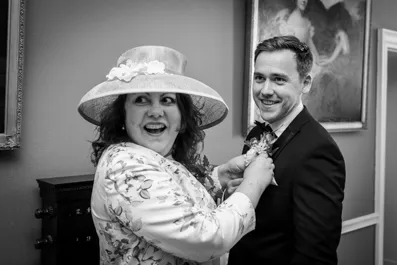 Smiling woman in a floral dress and hat adjusts the boutonniere on a young man's suit jacket in a room with framed paintings.