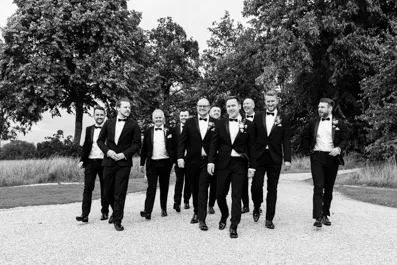 Group of eight men in tuxedos walking on a gravel path outdoors with trees in the background, smiling and engaged.
