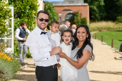 Smiling family of four outdoors on a sunny day with a man holding a baby boy and a woman holding a young girl.