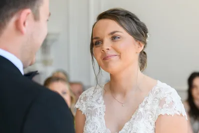 Bride with hair up wearing a white lace wedding dress smiling at groom during ceremony.