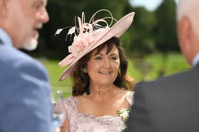 Smiling woman in a pink floral dress and matching wide-brimmed hat with decorative flowers, standing outdoors talking to two men.