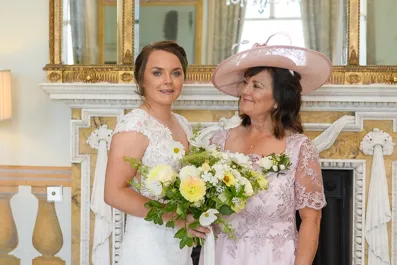 Bride in white lace dress holding bouquet standing next to woman in pink dress and matching hat in elegant room.
