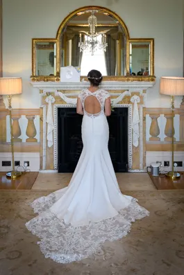 Bride standing with her back to the camera wearing a white wedding dress with lace train, in front of a decorated fireplace and mirror.