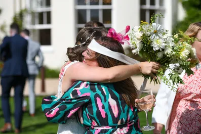 Two women hugging tightly outdoors at a celebration, one holding a bouquet and the other holding a wine glass.