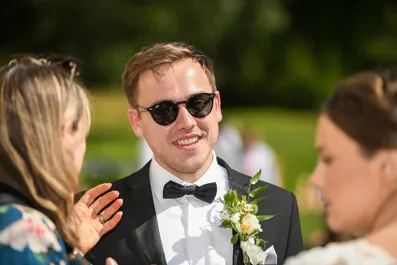 Man in a black tuxedo with a bow tie and boutonnière smiling while wearing sunglasses in an outdoor setting.