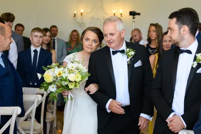Bride in white dress holding bouquet and linking arms with elderly man in tuxedo walking down aisle at wedding ceremony with guests watching.