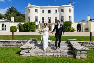 Bride and groom holding hands and walking down stone steps in front of a large white mansion on a sunny day.