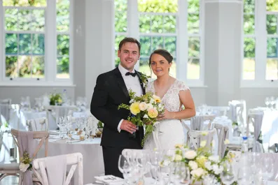 Bride in white lace wedding dress and groom in black tuxedo standing together holding a bouquet in a bright, elegantly decorated reception hall.