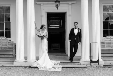 Black and white photo of a bride in a wedding gown holding a bouquet and a groom in a tuxedo standing on the steps of a building with large white columns.