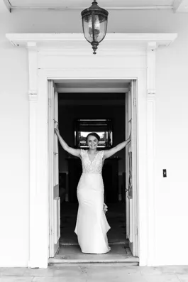 Bride in a lace wedding dress standing in a doorway with arms raised, smiling.
