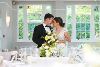 Bride and groom standing close together in a bright room with large windows, surrounded by wedding table settings with flowers and glassware.