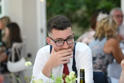 Man wearing glasses, a white shirt, and a red tie sitting at a table with hand covering his mouth, with blurred people and greenery in the background.