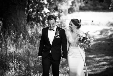 Black and white photo of a bride and groom walking hand in hand outdoors with trees and sunlight in the background.