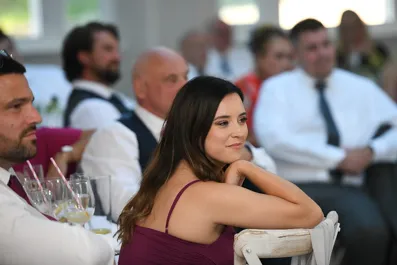 Woman in a purple dress sitting on a white chair, attentively listening at a formal event with other seated guests in the background.
