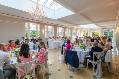 Guests seated at decorated round tables in a bright, elegant dining room with large windows and chandeliers.