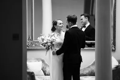 Bride and groom standing facing each other indoors with a bouquet and a large mirror behind them.