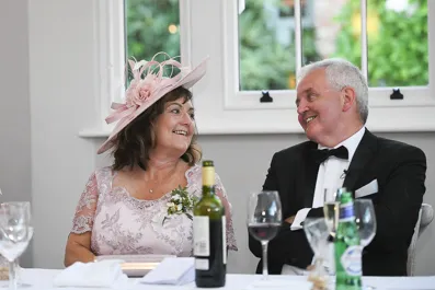 A smiling woman in a pink lace dress and hat sitting beside a man in a tuxedo at a table with drinks in front of a window.