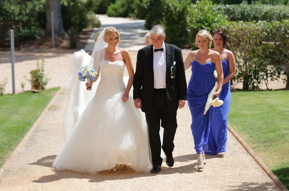 Bride in white wedding dress holding a bouquet walking down a path with a man in a tuxedo and two bridesmaids in purple dresses.