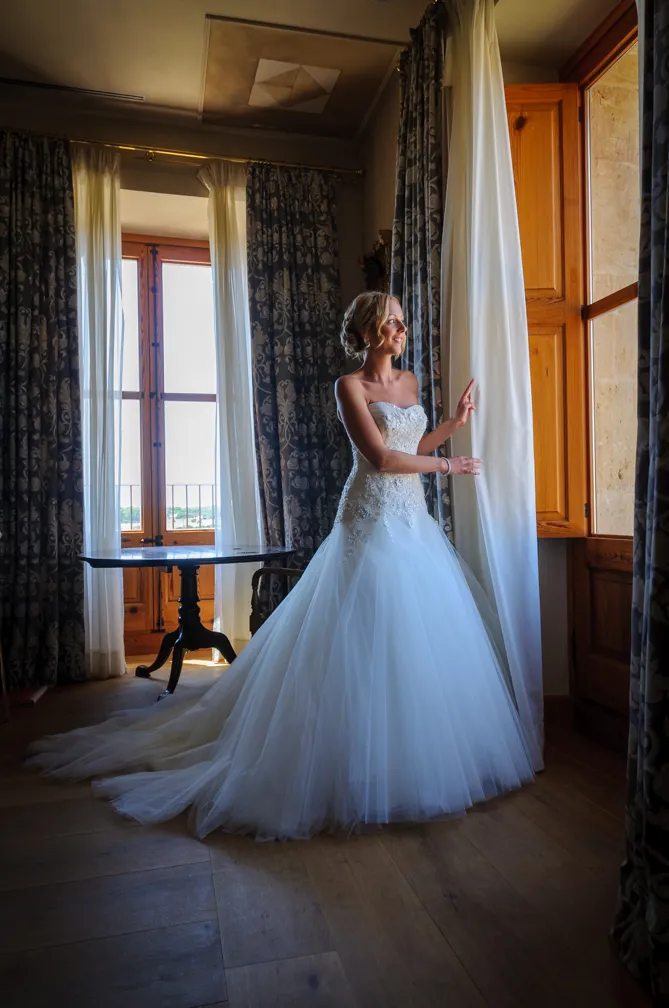 Bride in a white strapless wedding gown standing by a large wooden window with curtains, looking outside.