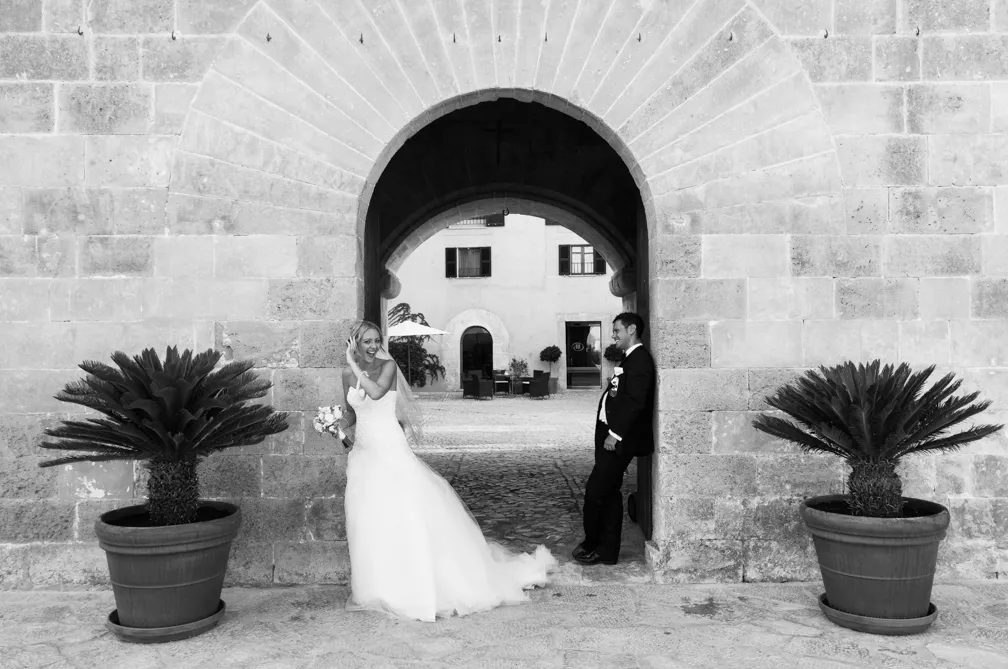 Bride in wedding dress and groom in suit posing playfully at stone archway entrance.