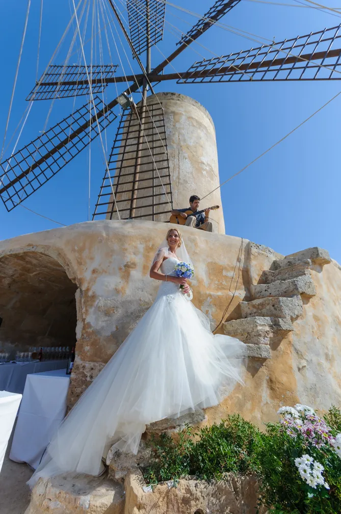 Bride in a flowing white gown holding a bouquet climbing stone steps with a man playing guitar sitting near a traditional windmill against a clear blue sky.