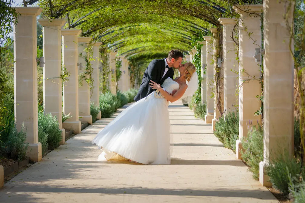Bride in white gown and groom in black tuxedo sharing a kiss under a green vine-covered archway.