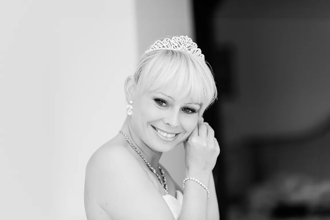 Smiling bride with short blonde hair wearing a tiara and earrings, adjusting an earring.
