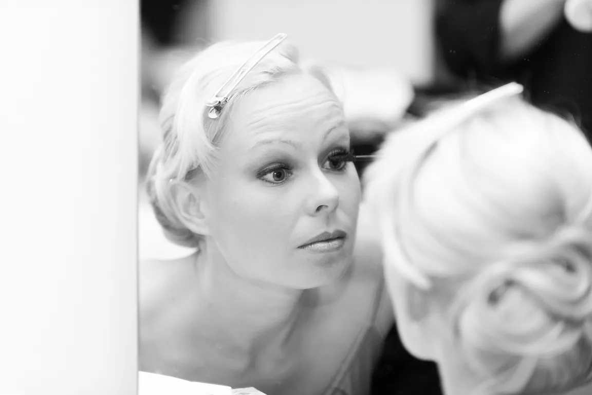 Woman with light hair applying mascara while looking into a mirror, hair pinned back.