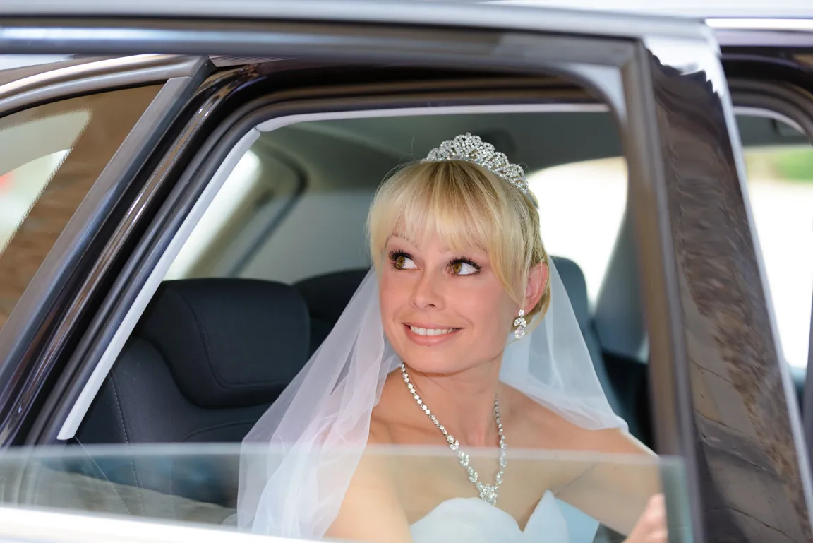 Smiling bride wearing a tiara and veil sitting inside a car.