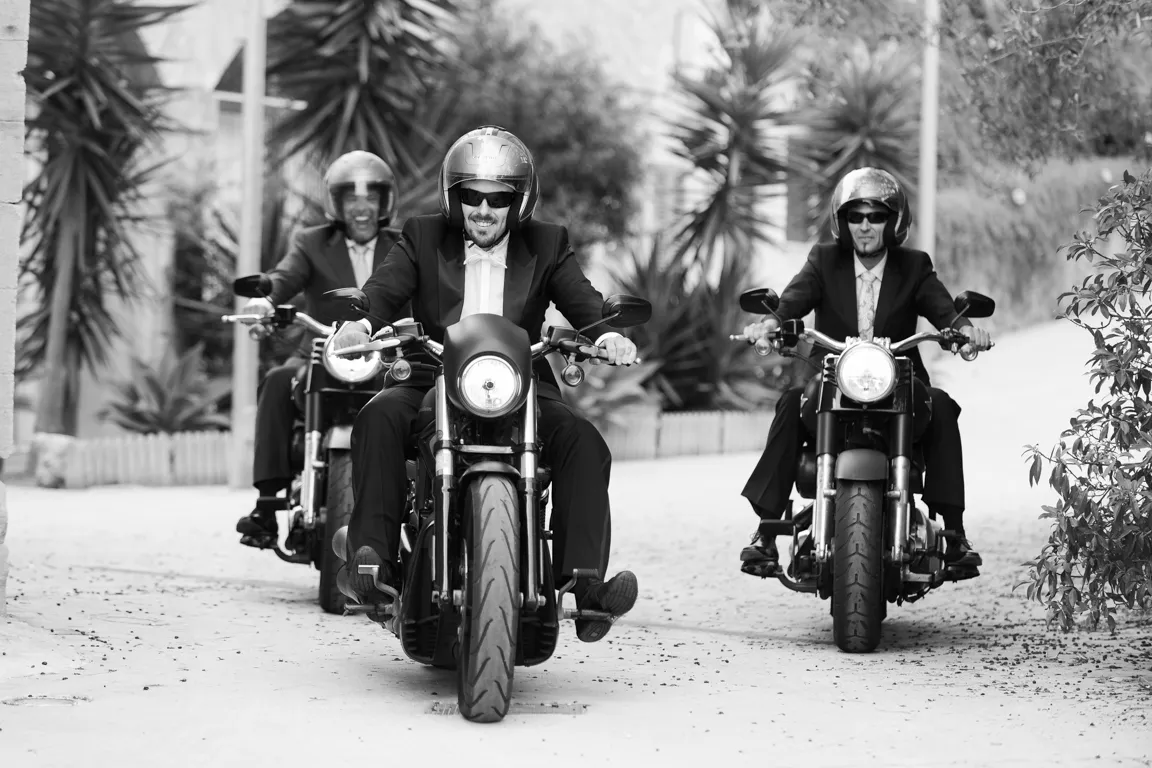 Three men in suits and helmets riding motorcycles on a street with plants in the background.