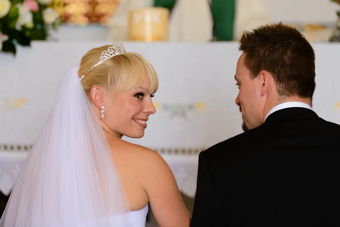 Smiling bride with tiara and veil looking back at the groom in a black suit inside a wedding venue.