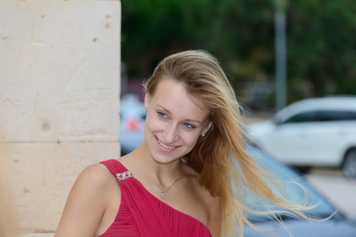 Young woman with long blonde hair wearing a red one-shoulder dress, smiling and looking to her left outdoors.