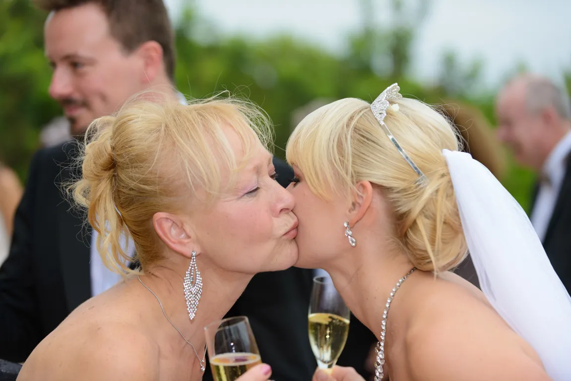 Bride and an older woman sharing a cheek kiss while holding champagne glasses at an outdoor event.