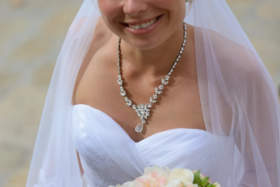 Close-up of a smiling bride wearing a white strapless wedding dress, a veil, and a sparkling crystal necklace, holding a bouquet.