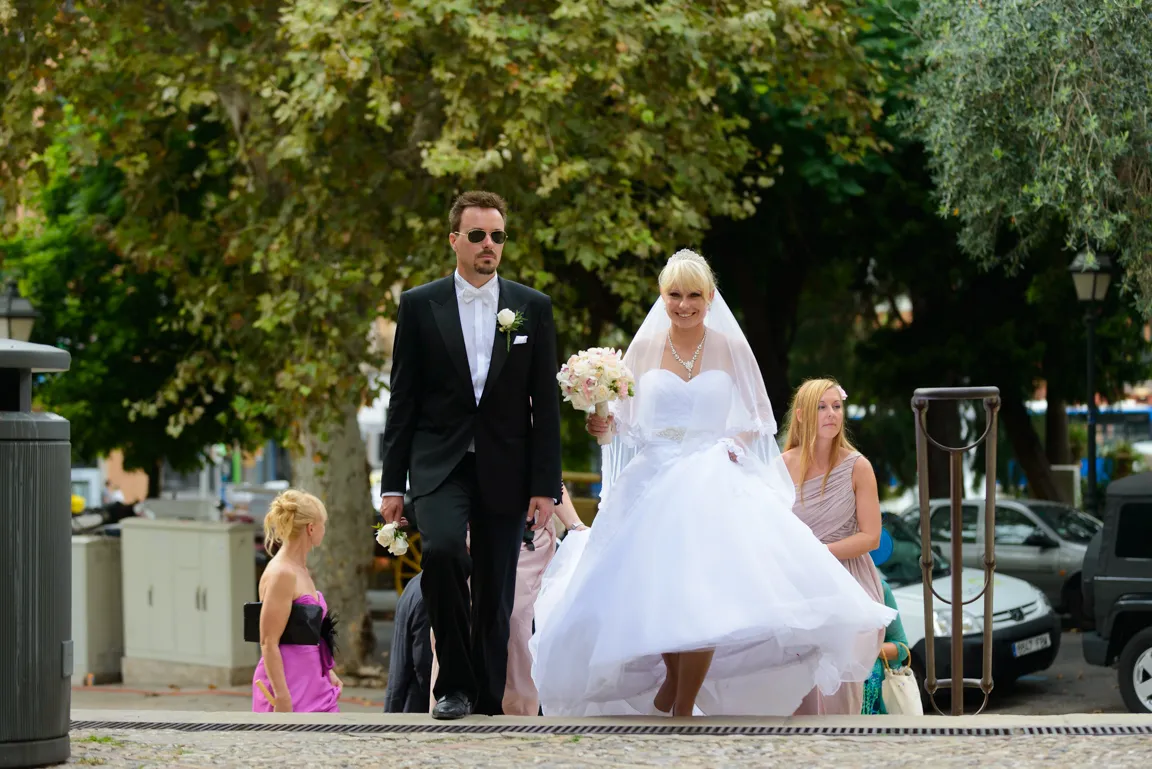 Bride in a white wedding dress holding a bouquet, walking beside a groom in a black tuxedo and sunglasses, with bridesmaids behind them outdoors.